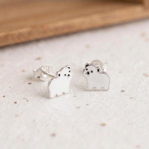 Puede incluir: Un par de pendientes de botón con forma de oso polar. Cada pendiente presenta un relleno de esmalte blanco en el cuerpo, con detalles en esmalte negro para los ojos y la nariz. Los postes de plata son visibles y marcados con 925.
