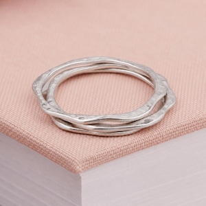 Op de afbeelding: Een set van drie zilverkleurige ringen met een gehamerde, onregelmatige textuur. De ringen zijn op elkaar gestapeld en hebben een golvende, organische vorm met een matte afwerking. Elke ring heeft een consistente dikte en een ongelijk profiel.
