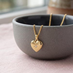 May include: A gold-toned heart-shaped pendant necklace featuring a small, centered starburst engraving with a single embedded clear stone. The pendant hangs from a delicate gold-toned chain, showcasing a polished, smooth metallic finish throughout.