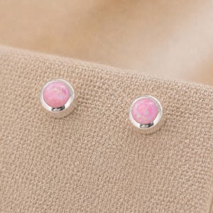 May include: A pair of stud earrings featuring round, cabochon-cut pink opal stones set in polished silver-toned metal bezels. The stones display a smooth, domed surface with subtle color variations, securely held within the circular metallic frames.