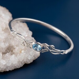 May include: A silver bangle bracelet featuring an oval-shaped light blue gemstone set in the center. The metal band is designed with intricate, flowing leaf-like filigree patterns extending from both sides of the central stone setting.