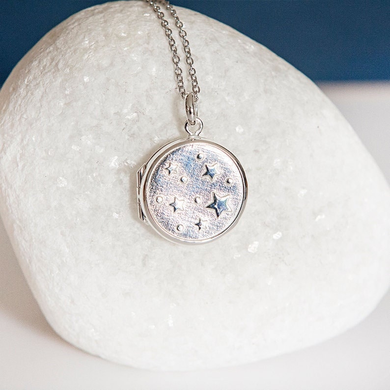 Puede incluir: Collar con colgante plateado y cadena fina. El colgante redondo presenta un dise&ntilde;o celestial de estrellas y puntos. El colgante est&aacute; cerrado, lo que sugiere que puede contener una peque&ntilde;a foto o recuerdo. El collar se muestra sobre una superficie blanca.