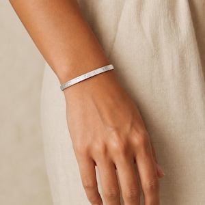 May include: A slim, silver-toned metal bangle bracelet featuring a repeating pattern of small, etched star-like symbols along its outer surface. The bracelet has a smooth, polished finish and a thin, uniform band design that encircles the wrist.