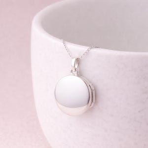 Puede incluir: Un colgante de medall&oacute;n redondo plateado sobre una delicada cadena. El medall&oacute;n es liso y reflectante, con un dise&ntilde;o de bisagra. El collar se muestra sobre un fondo blanco.