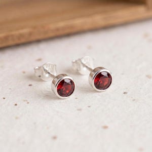 May include: A pair of stud earrings featuring round, faceted deep red garnet gemstones set in polished silver bezels. Each earring includes a silver post and a matching butterfly clutch back, showcasing a simple and clean design.