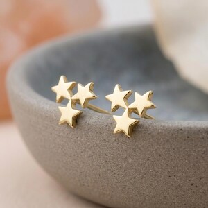 May include: A pair of gold stud earrings, each featuring a cluster of three small, polished five-pointed stars arranged in a triangular formation. The earrings have a smooth, reflective metallic finish and include thin posts extending from the back.