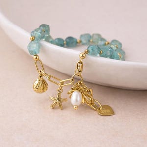 Puede incluir: Una pulsera con dijes dorada con cuentas azul claro. La pulsera incluye un dije de concha, estrella de mar, perla y corazón. La pulsera se muestra sobre una superficie blanca y curvada.