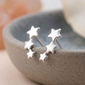 Puede incluir: Un par de pendientes de botón en tono plateado, cada uno con tres estrellas conectadas de diferentes tamaños dispuestas en línea vertical. Las estrellas tienen un acabado pulido y liso que refleja la luz, e incluyen un cierre de poste estándar.