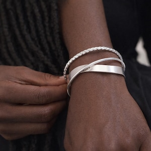 May include: Three silver bracelets are displayed on a wrist. The bracelets include a twisted rope design, a thin band, and a wide, flat band that crosses over the other two. The bracelets are made of a shiny, reflective metal.