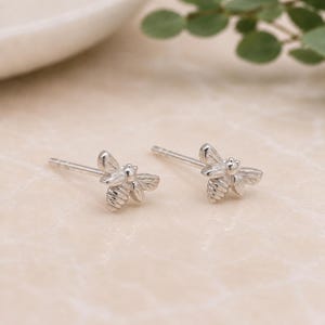 Peut inclure: Une paire de boucles d'oreilles en forme d'abeille en argent. Chaque boucle d'oreille présente un motif d'abeille détaillé avec des ailes et des segments de corps. Les boucles d'oreilles sont posées sur un fond doux et neutre.