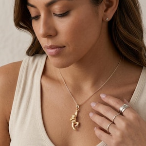 May include: A gold-toned necklace featuring a small, detailed monkey pendant suspended from a delicate chain. The monkey is depicted in a hanging position with textured fur details. The necklace is worn with two simple silver-toned rings on the fingers.