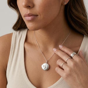 May include: A round silver locket pendant featuring an embossed bee design, suspended from a delicate silver chain. The jewelry set includes three silver rings worn on the fingers, showcasing a polished metallic finish and a circular locket shape.