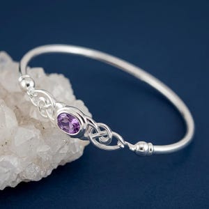 May include: A silver bangle bracelet featuring a central oval-cut purple amethyst gemstone. The metal band displays intricate Celtic-style knotwork detailing on both sides of the stone, finished with smooth, rounded silver end caps.