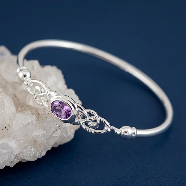 May include: A silver bangle bracelet featuring a central oval-cut purple amethyst gemstone. The metal band displays intricate Celtic-style knotwork detailing on both sides of the stone, finished with smooth, rounded silver end caps.
