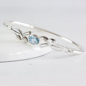 May include: A silver bangle bracelet featuring a central oval-cut light blue gemstone. The metal band is designed with symmetrical leaf-like motifs extending from both sides of the stone setting, creating a continuous polished metallic finish.