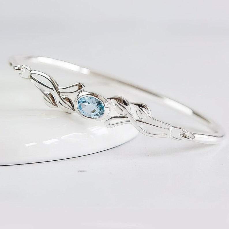 May include: A silver bangle bracelet featuring a central oval-cut light blue gemstone. The metal band is designed with symmetrical leaf-like motifs extending from both sides of the stone setting, creating a continuous polished metallic finish.