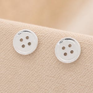 May include: Two circular, silver-toned buttons with a polished, reflective finish. Each button features a recessed center with four evenly spaced holes for thread, set against a textured, light beige fabric background.