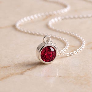 May include: A silver necklace featuring a round, faceted, deep red gemstone pendant set in a polished silver bezel. The pendant is attached to a delicate silver chain with small, circular links that extend outward from the central setting.