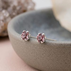 May include: A pair of silver stud earrings featuring round, faceted, light pink stones held in place by six-prong settings. The earrings have a metallic post back and are displayed with the stones facing forward, showcasing the clear, reflective surfaces.