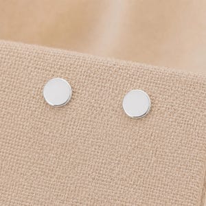 Pode incluir: Um par de pequenos brincos de botão circulares em prata com uma superfície lisa, polida e refletora. Os brincos são simples, redondos e metálicos, apresentando uma face plana que capta a luz, apresentados como um conjunto de duas peças.