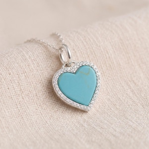 May include: A silver heart-shaped pendant necklace featuring a smooth, light blue turquoise stone inlay at the centre. The heart is framed by a border of small, clear, sparkling stones and hangs from a delicate silver chain link.