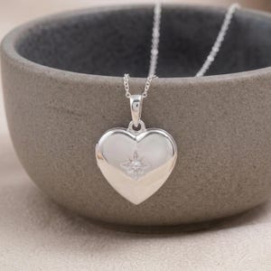 May include: A polished silver heart-shaped locket pendant suspended from a delicate silver chain. The center of the heart features a small, engraved starburst design with a single, tiny embedded clear stone at its focal point.