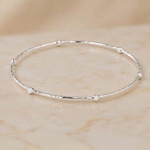 May include: A thin, circular silver bangle bracelet featuring a hammered, textured surface. The bangle is accented with five small, evenly spaced round white opal stones set into the metal band, creating a delicate and reflective appearance.