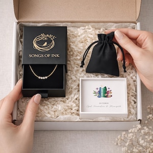 May include: A gift box containing a black sliding jewelry box with a gold bird logo and the text SONGS OF INK, a delicate gold necklace with four small round stones, a black drawstring pouch, and a white card featuring a crystal illustration.