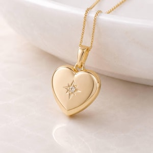 Puede incluir: Un collar con colgante en forma de corazón dorado. El colgante presenta un diseño de estrella con un pequeño diamante en el centro. El collar tiene una cadena fina y se muestra sobre una superficie blanca.