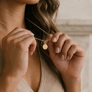 May include: A gold-toned necklace featuring a delicate chain and a small, circular pendant. The pendant has a textured, engraved surface with a subtle botanical or floral design. The necklace rests against the chest, showcasing the metallic finish.