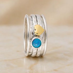 May include: A silver spinner ring featuring a hammered texture on the central band. The ring is adorned with a small, smooth gold-toned heart charm and a circular, vibrant blue opal-like stone set in a polished silver bezel on the outer surface.