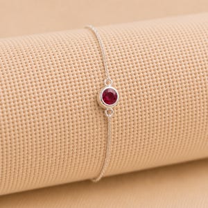 Puede incluir: Una delicada pulsera de cadena de plata con una piedra preciosa roja redonda engastada en el centro. La pulsera está construida con una fina cadena de eslabones de plata pulida que se conecta al engaste metálico circular que sujeta la piedra.
