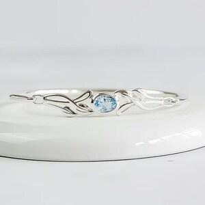 May include: A silver bangle bracelet featuring a central oval-cut light blue gemstone set in a bezel. The metal band displays a polished finish with symmetrical, flowing, leaf-like decorative elements extending from the center stone.