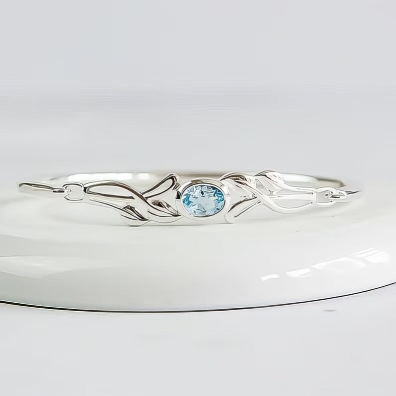 May include: A silver bangle bracelet featuring a central oval-cut light blue gemstone set in a bezel. The metal band displays a polished finish with symmetrical, flowing, leaf-like decorative elements extending from the center stone.