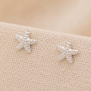 May include: A pair of silver-toned stud earrings shaped like five-pointed starfish. Each earring features a textured surface with small, raised bumps across the entire body, mimicking the natural appearance of a sea creature in a metallic finish.