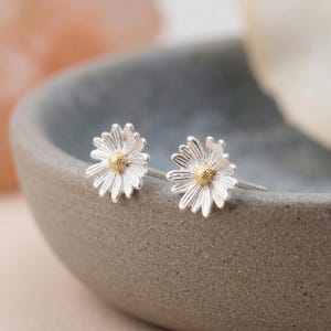 Puede incluir: Un par de pendientes de botón con forma de flores de margarita. Cada pendiente presenta pétalos de tono plateado que irradian desde un centro de tono dorado texturizado. El diseño es consistente en ambas piezas, mostrando un motivo floral.