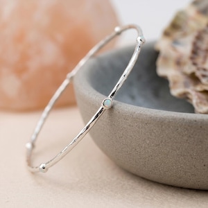 May include: A thin, silver-toned bangle bracelet featuring a hammered, textured surface. The circular band is accented with small, evenly spaced, bezel-set round opals that display subtle iridescent flashes of colour against the polished metal.