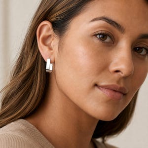 May include: A small, polished silver hoop earring with a rectangular, open-link shape. The earring features a smooth, reflective metallic surface and a secure hinged closure, worn on the earlobe to provide a clean, geometric aesthetic.