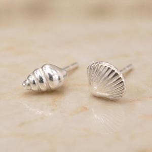 May include: A pair of silver stud earrings featuring a polished spiral conch shell design and a textured fan-shaped scallop shell design. Each earring has a metal post backing, showcasing a metallic finish with detailed ridges and smooth curves.