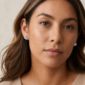 May include: A pair of small, silver-toned stud earrings shaped like seashells. Each earring features a fan-like design with delicate, vertical ridges and a dark, iridescent inlay that catches the light, providing a subtle contrast to the polished metal edges.