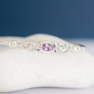 May include: A silver bangle bracelet featuring a central oval-shaped light purple gemstone. The metalwork displays intricate, interconnected loops on both sides of the stone, flanked by smooth, polished silver spheres on the band.