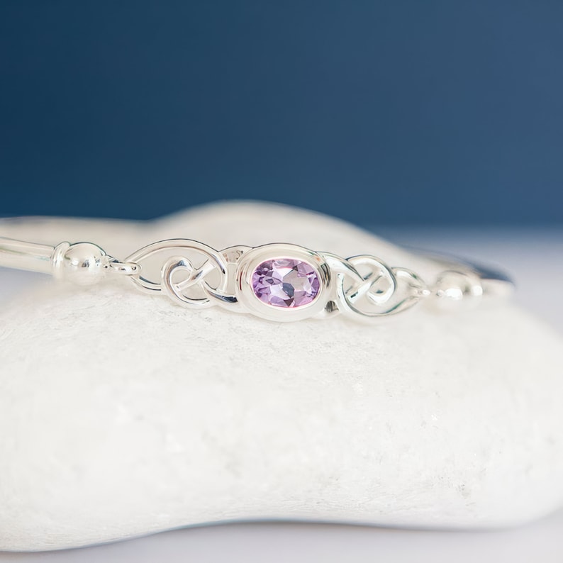 May include: A silver bangle bracelet featuring a central oval-shaped light purple gemstone. The metalwork displays intricate, interconnected loops on both sides of the stone, flanked by smooth, polished silver spheres on the band.