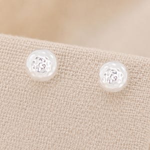 May include: A pair of round silver stud earrings featuring a textured, hammered surface finish. Each earring has a polished, reflective metallic appearance with small, irregular indentations across the dome-shaped front, creating a subtle light-catching effect.