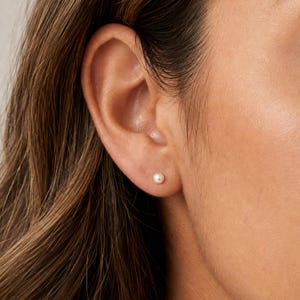 May include: A small, round, white pearl stud earring is centered in the earlobe. The pearl has a smooth, lustrous surface and a classic spherical shape, providing a simple and clean appearance against the skin of the ear.