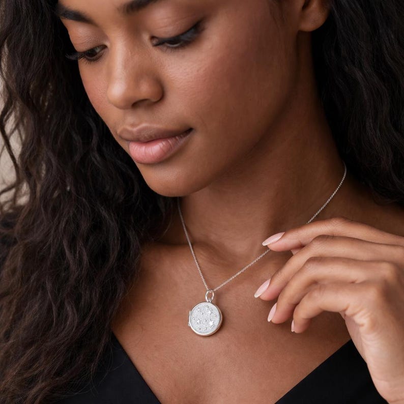 Puede incluir: Un collar de plata con un colgante redondo. El colgante tiene un fondo blanco con peque&ntilde;os detalles brillantes. El collar lo lleva una persona que viste un top negro.