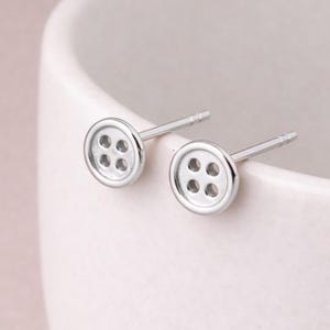 Op de afbeelding: Een paar zilveren knoopvormige oorstekers. Elke oorbel heeft een cirkelvormig ontwerp met vier kleine gaatjes, die lijken op een klassieke knoop. Op een zachte, neutrale achtergrond.