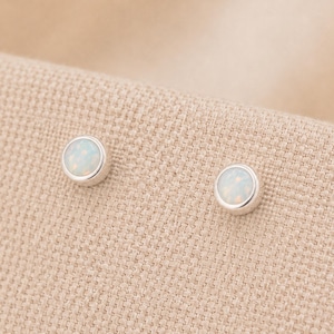 May include: A pair of round stud earrings featuring smooth, bezel-set white opal cabochons. Each earring has a polished silver-tone metal rim surrounding the iridescent, milky white stone, creating a simple and clean circular design for the ears.