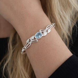 May include: A silver bracelet featuring a central oval-cut light blue gemstone. The metal band is designed with intricate, symmetrical leaf-like motifs extending from the center stone, creating a continuous, polished metallic structure.