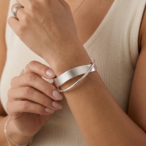 Pode incluir: Uma pulseira de prata com uma banda larga e plana de acabamento escovado que se sobrepõe a uma banda de fio mais fina, polida e arredondada. A pulseira é usada no pulso, com um anel de prata a condizer visível no dedo indicador.
