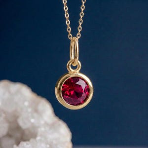May include: A gold pendant necklace featuring a round, faceted deep red gemstone set in a polished gold bezel. The pendant hangs from a delicate gold chain, showcasing a classic circular design with a vibrant, reflective crimson stone centre.